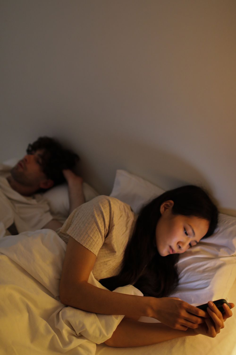 Kvinde ligger i sengen og kigger på sin mobil, mens hendes partner sover ved siden af