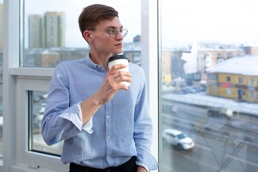 Mand med briller står ved et vindue med en kop kaffe og ser eftertænksom ud